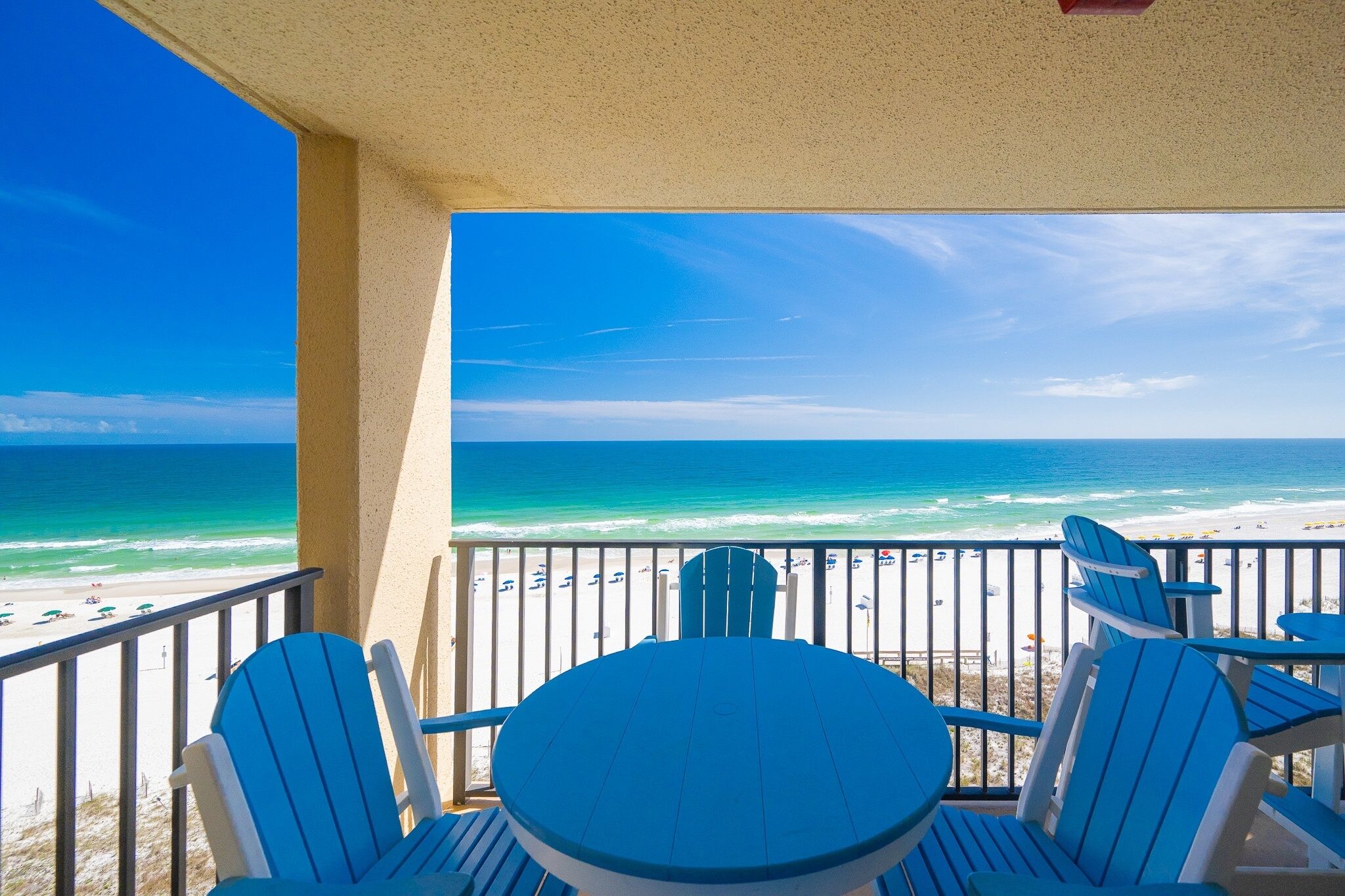 A balcony with blue Adirondack chairs and a table, overlooking a beach with umbrellas and people.