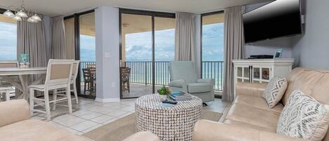 A spacious living room with a large window overlooking the ocean, a ceiling fan, and a flat-screen TV mounted on the wall.