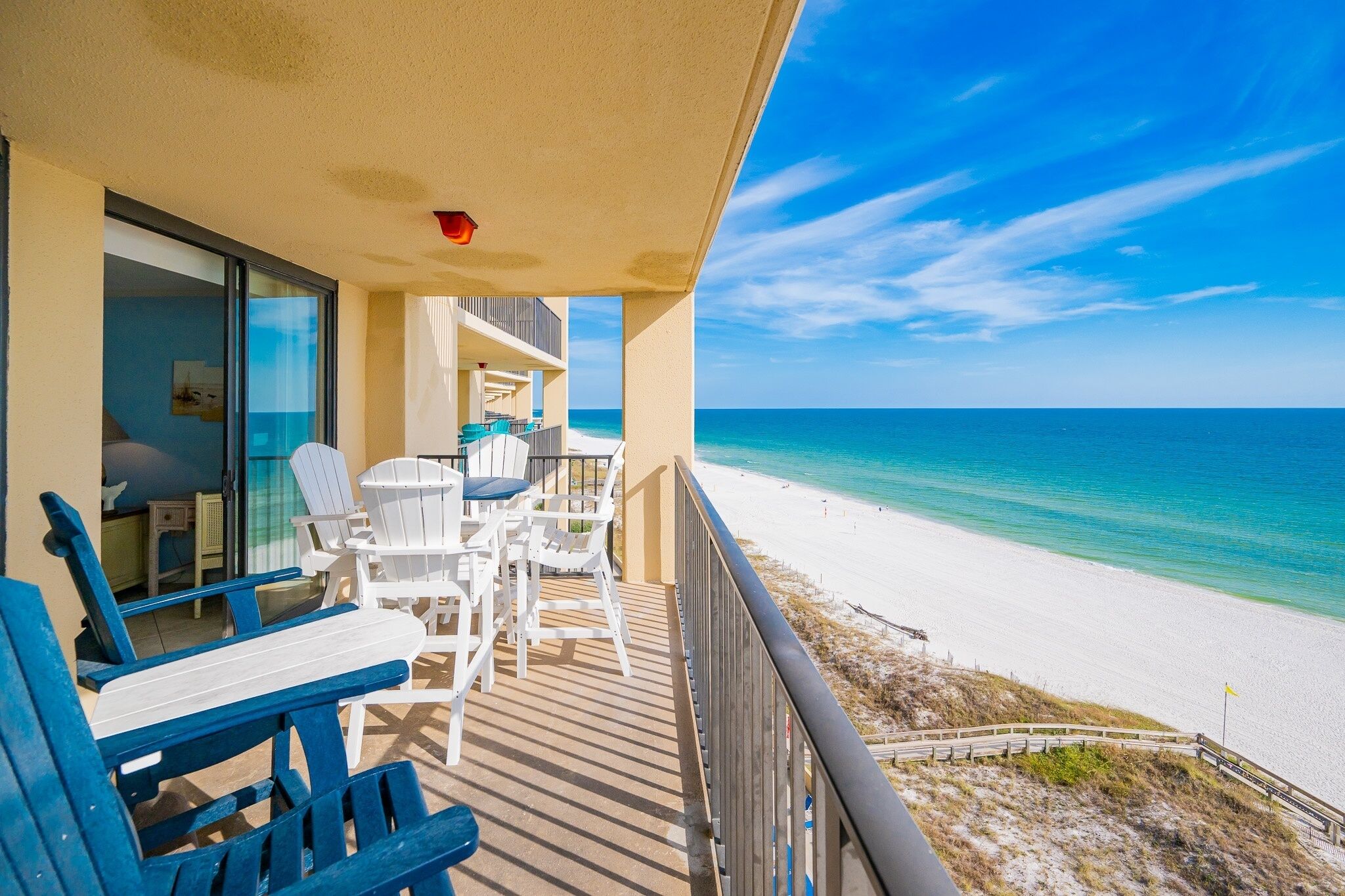 Un balcon avec des chaises blanches et une table, donnant sur une plage aux eaux d’un bleu limpide et au rivage sablonneux.