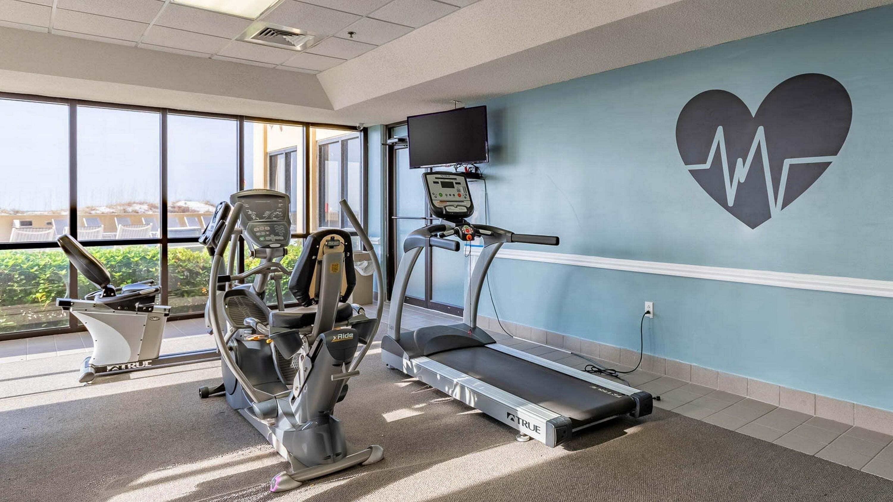 Une salle de remise en forme équipée de tapis de course, d’un vélo elliptique et d’un vélo d’appartement, avec de grandes fenêtres et une décoration murale en forme de cœur.