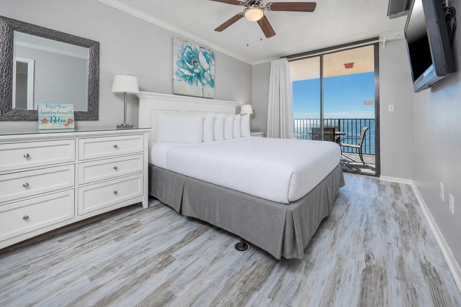 A bedroom with a bed, a dresser, a ceiling fan, a painting, and a balcony with a view.