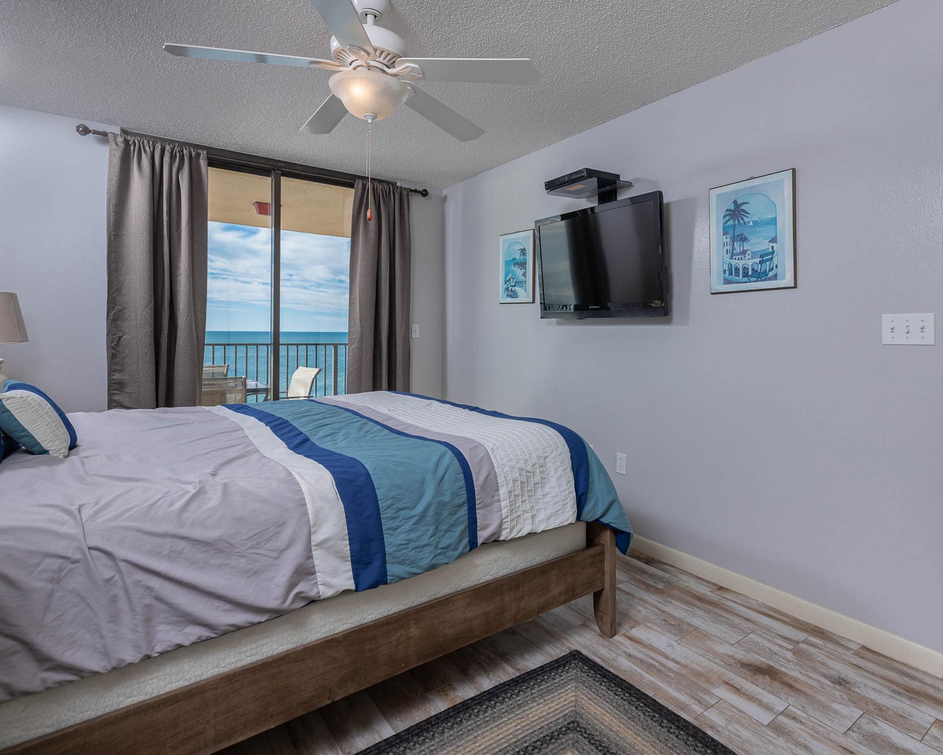 Une chambre à coucher avec un lit, un ventilateur de plafond, une télévision et un balcon offrant une vue sur la mer.