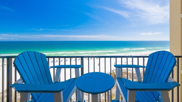 A balcony with two blue Adirondack chairs and a small round table, overlooking a beach with clear blue water and a sandy shore. - Phoenix V by Brett/Robinson Vacations (Orange Beach)
