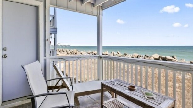 A covered balcony with a view of the beach, a chair, a table, and a door. - A Sea Escape 1 Bedroom Villa (Carolina Beach)