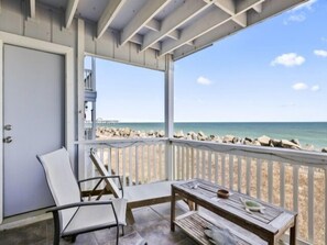 A covered balcony with a view of the beach, a chair, a table, and a door. - A Sea Escape 1 Bedroom Villa (Carolina Beach)