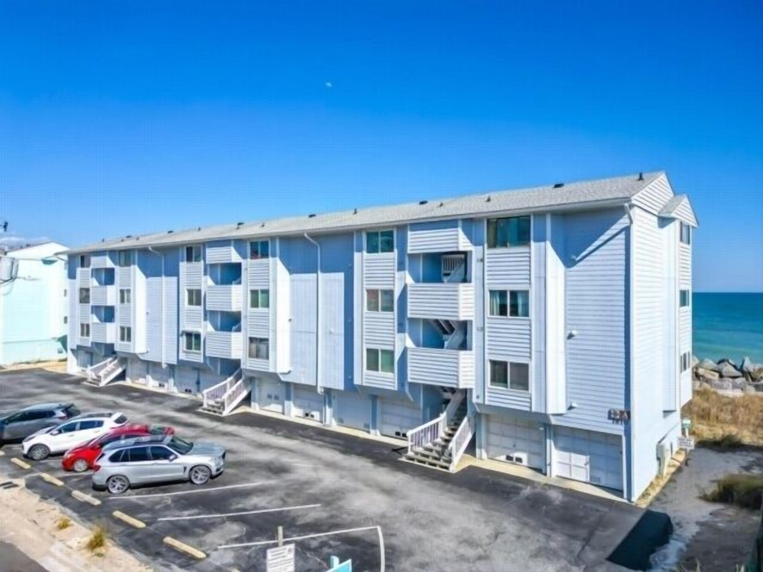 A multi-story motel with a row of rooms, a parking lot with several cars, and a view of the ocean.