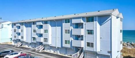 A multi-story motel with a row of rooms, a parking lot with several cars, and a view of the ocean.