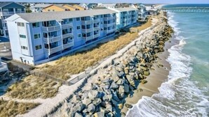 A coastal hotel with multiple floors and balconies, situated next to a rocky shoreline with waves crashing onto the beach. - A Sea Escape 1 Bedroom Villa (Carolina Beach)