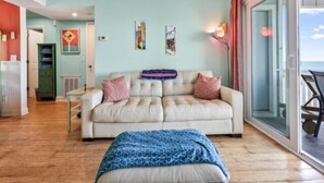 A living room with a beige sofa, a blue patterned ottoman, and a glass door leading to a balcony. - A Sea Escape 1 Bedroom Villa (Carolina Beach)