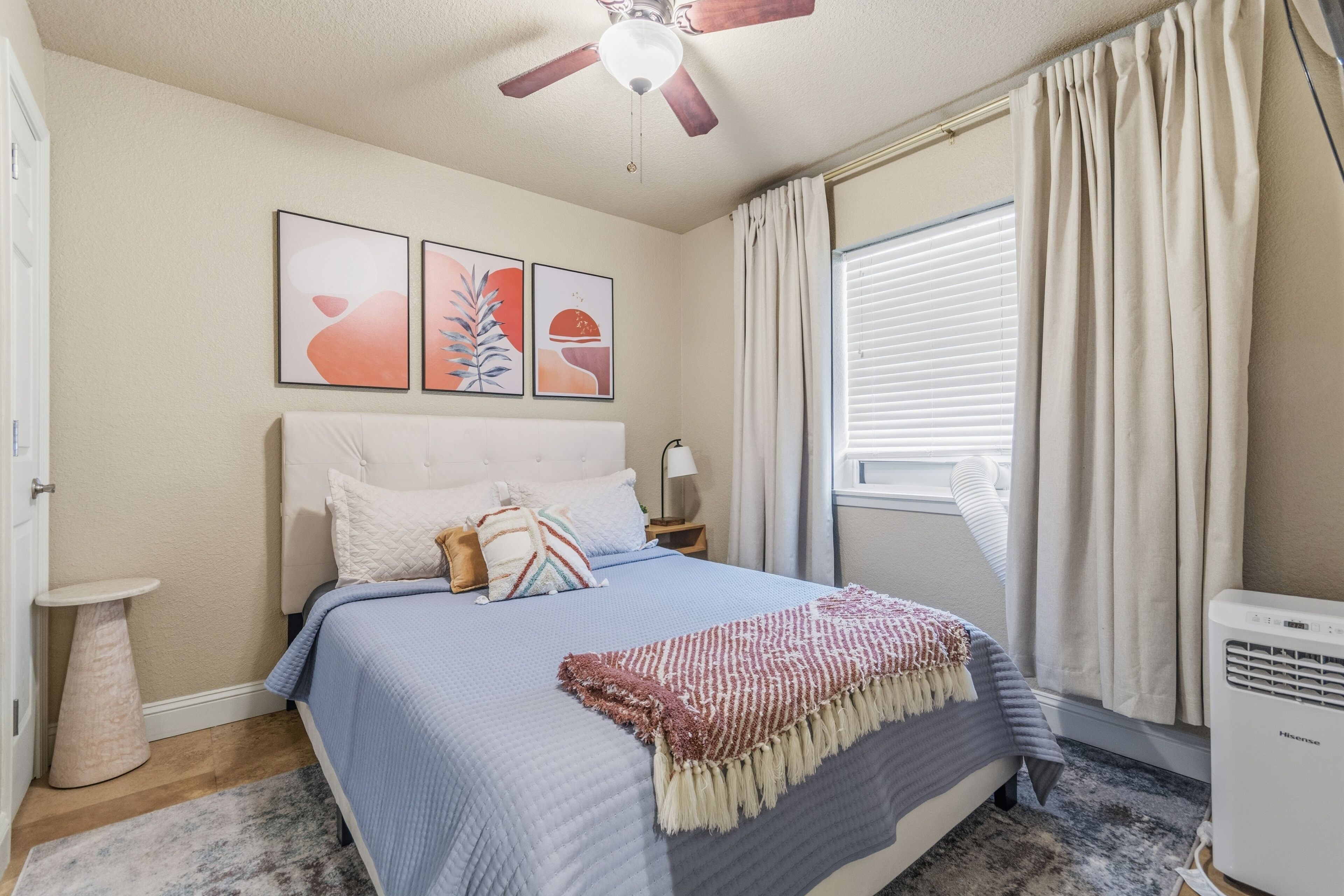 A bedroom with a bed, a ceiling fan, a window with curtains, and artwork on the wall.
