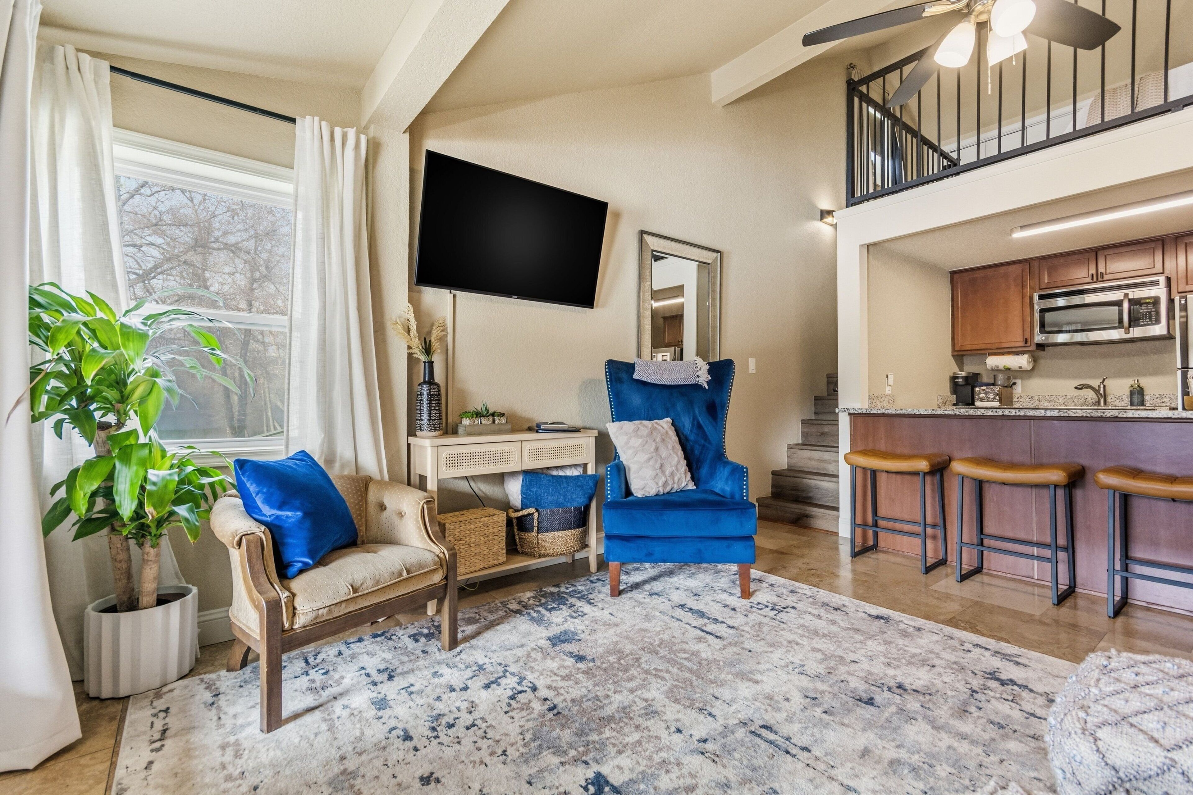 Een woonkamer met een bank, een blauwe fauteuil, een salontafel, een televisie, een spiegel, en een keuken met een barkruk.