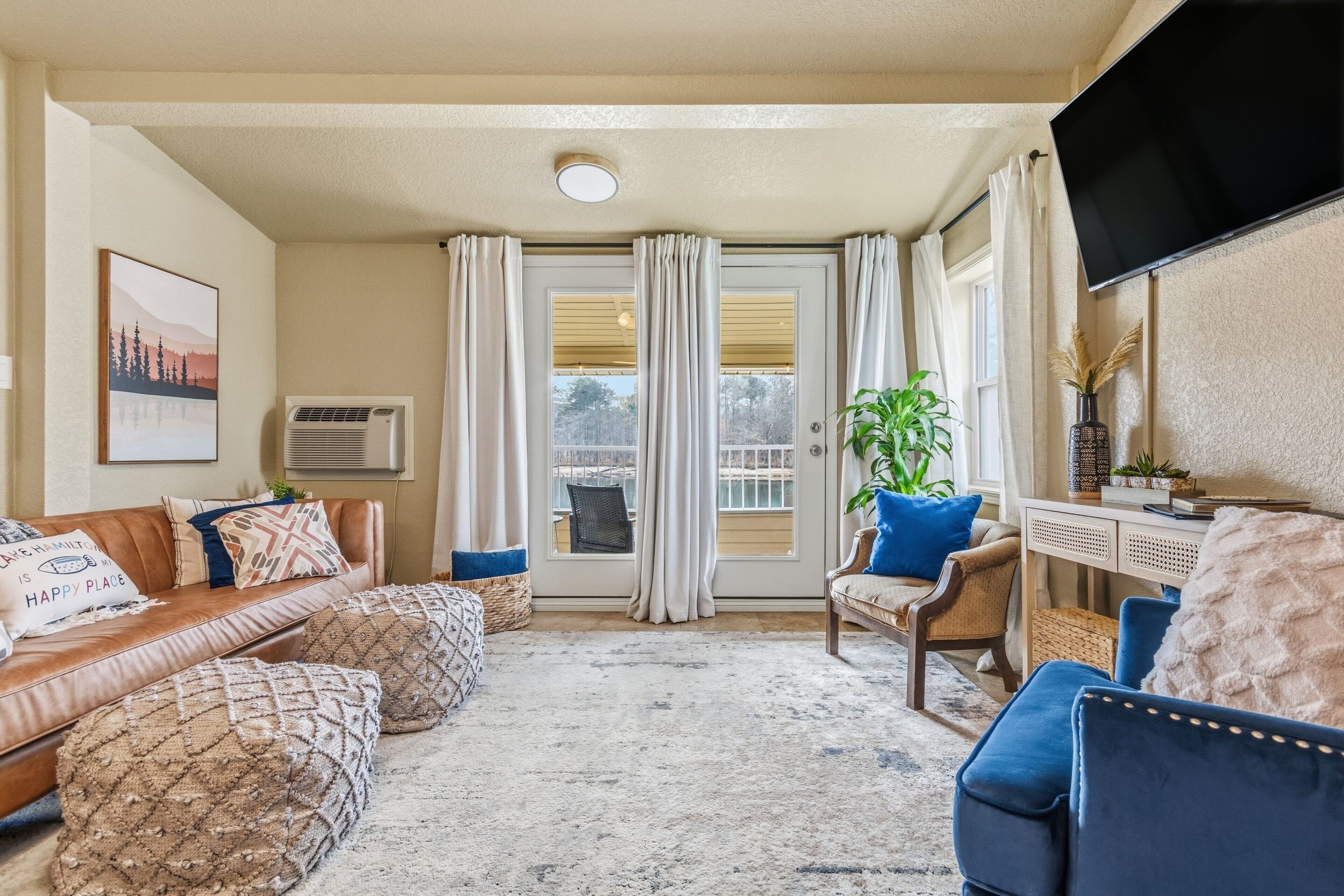 Un salon avec un canapé en cuir marron, des fauteuils bleus et un tapis beige. Il y a une grande fenêtre avec des rideaux, un téléviseur à écran plat fixé au mur et une plante sur le rebord de la fenêtre.