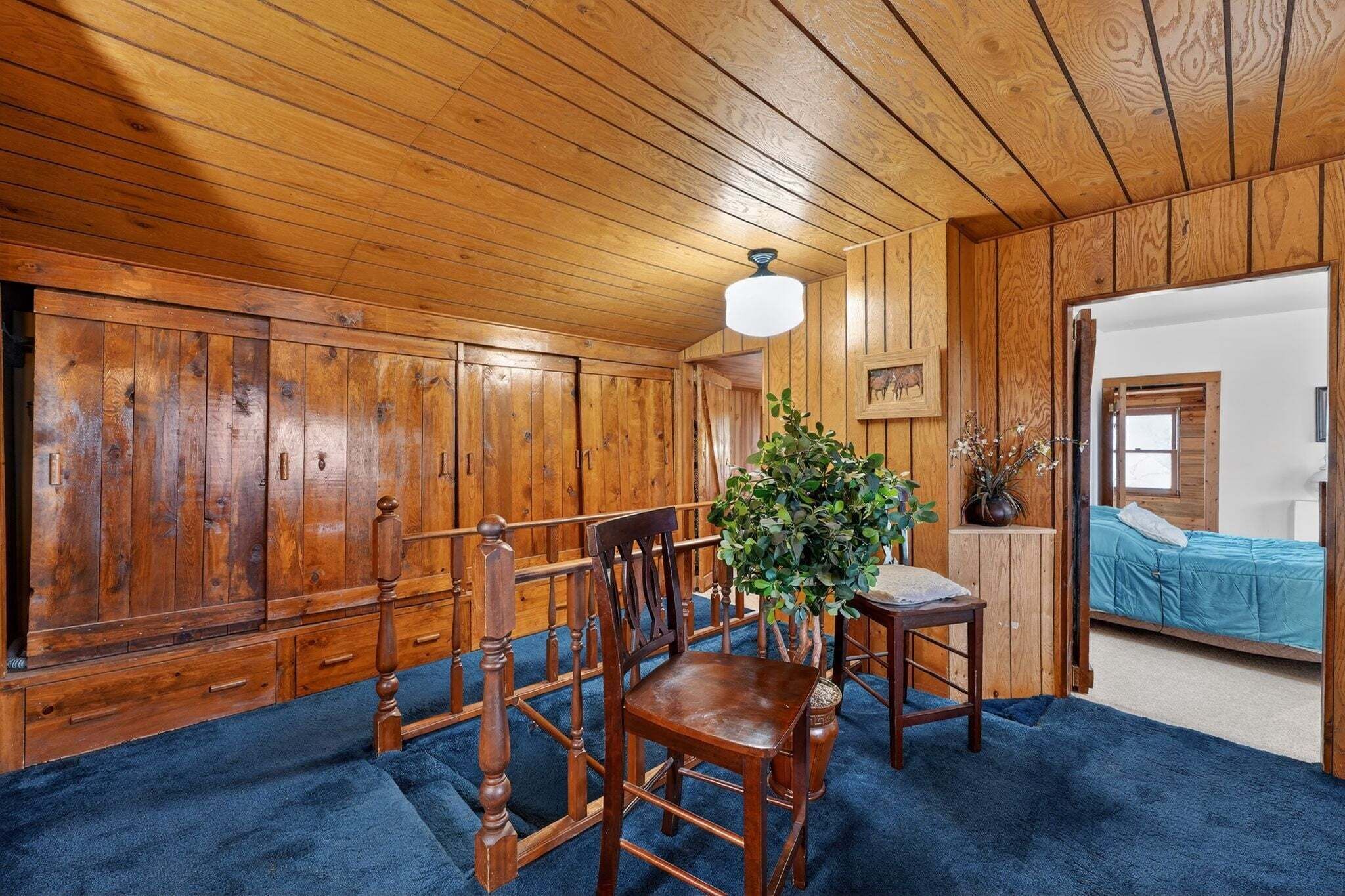 A room with wooden walls and ceiling, a blue carpet, a wooden dining table with chairs, a potted plant, and a bed with a blue bedspread.