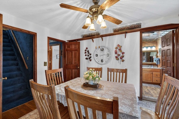 A dining area with a wooden table, chairs, a ceiling fan, and a clock on the wall. - Rustic Shadows - Sioux Falls Escape, Firepit, Pet Friendly! (Chancellor)