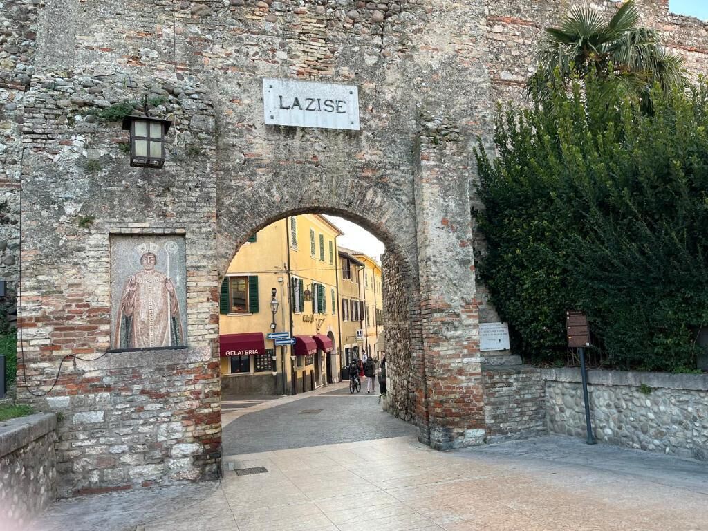 En portal med en skylt som visar texten ”LAZISE”, som leder in på en gata med byggnader och en glassbar.