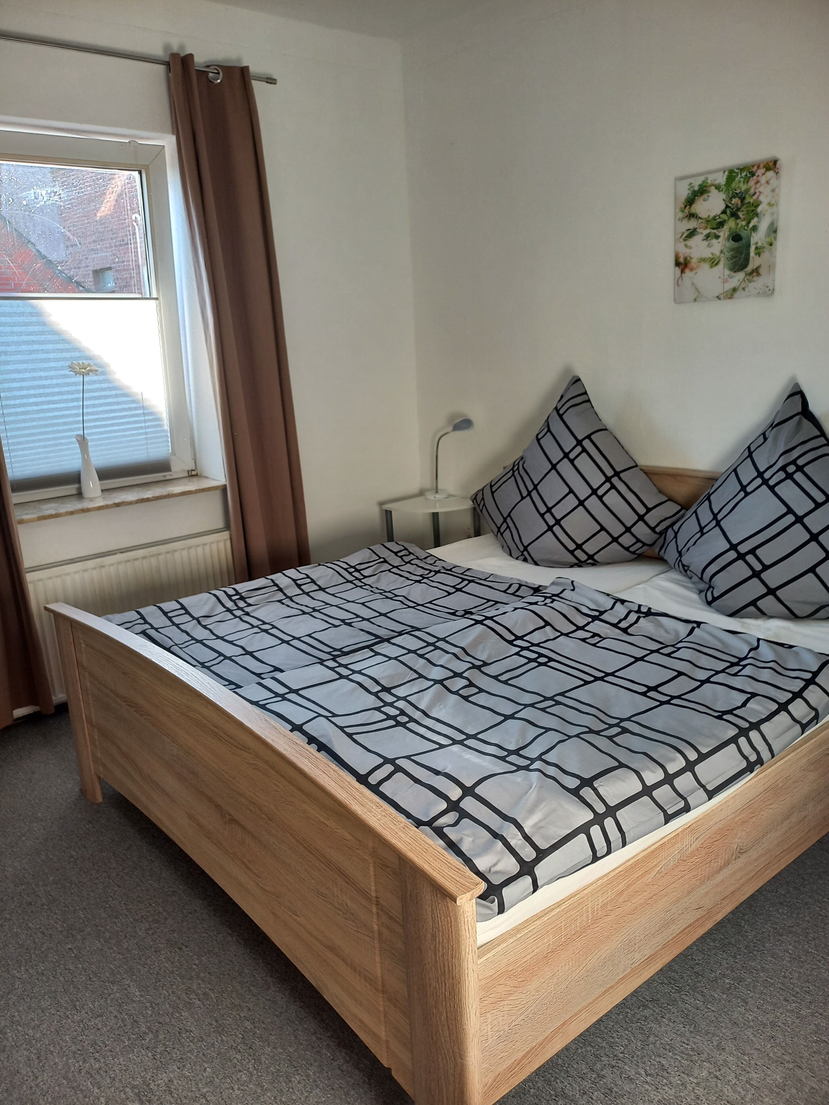 A bedroom with a wooden bed, a window with curtains, a bedside table, and a wall-mounted lamp.