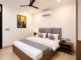 A hotel room with a bed, a ceiling fan, an air conditioning unit, a bedside table with a lamp, and a painting on the wall.