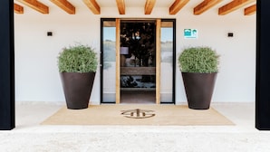 A modern building entrance with a wooden ceiling, large potted plants, and a welcome mat with a logo. - FINCA LOS COLLADOS DE SAN MIGUEL (Villamanrique)