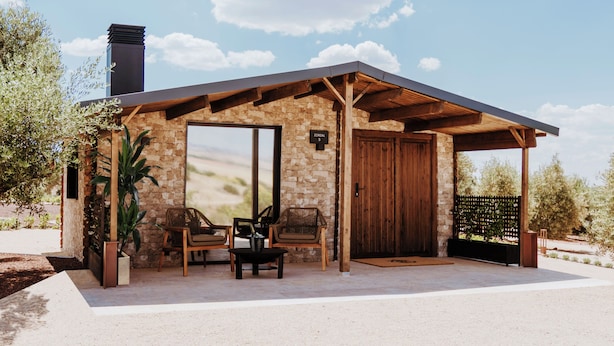 A modern house with a stone exterior, a wooden roof, and a large door. There are two chairs and a table outside. - FINCA LOS COLLADOS DE SAN MIGUEL (Villamanrique)