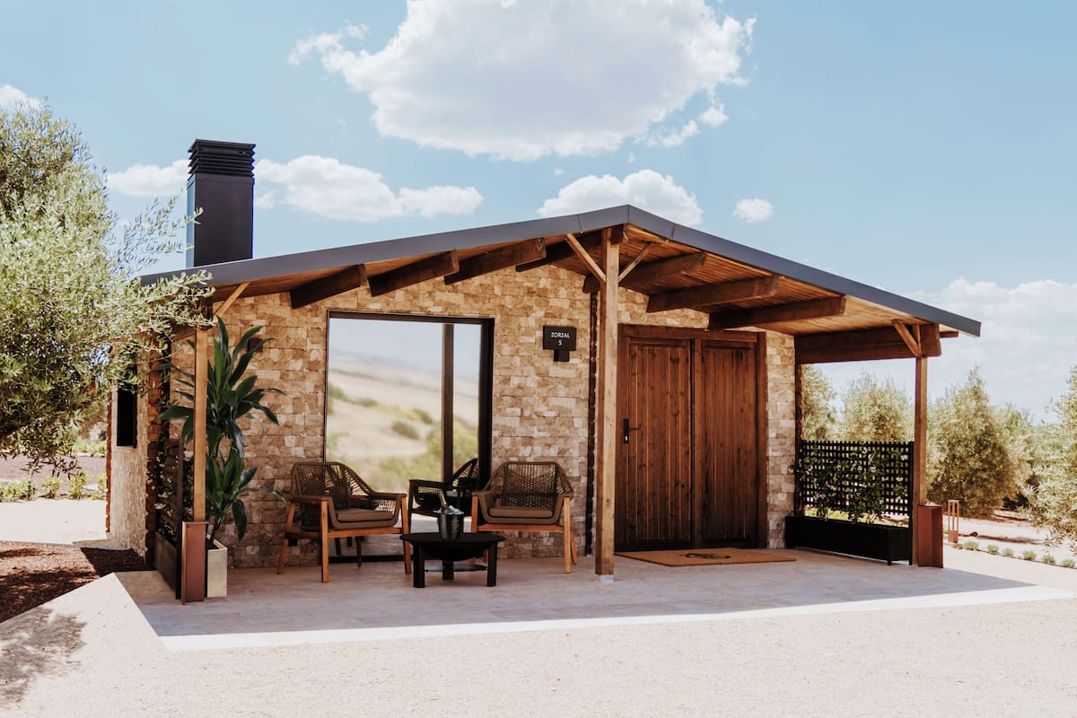 Ein modernes Haus mit Natursteinfassade, Holzdach und einer großen Tür. Vor der Tür stehen zwei Stühle und ein Tisch.