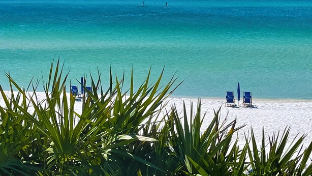 Een strand met helder turquoise water, twee mensen die paddleboarden, en meerdere strandstoelen op het zand.