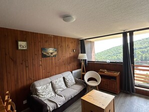 Ein Zimmer mit Holzwand, einer Couch, einem kleinen Tisch, einer Lampe und Blick auf einen bewaldeten Hügel.