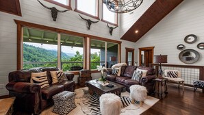 A spacious living room with a large window offering a mountain view, leather sofas with zebra-patterned cushions, and a rustic wooden coffee table. - Gatlinburg Splash-Home Theater-Heated Indoor Pool-Game Room! (Gatlinburg)