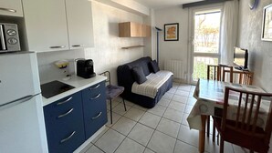 Una cucina funzionale con frigorifero, fornello e macchina del caffè. Un divano e un tavolo da pranzo sono posizionati vicino a una finestra, con una sedia e una TV su un mobile.