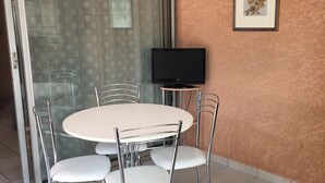 A dining area with a round table and four chairs, a television on a stand, and a patterned curtain. - Ground Floor with Private Parking for 4 (Argelès-sur-Mer)