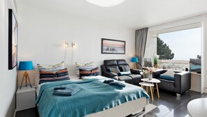 A modern hotel room with a bed, a sofa, a chair, a small table, and a large window. - Apartment with Sea View, Sauna, and High-Quality Furnishings (Sierksdorf)