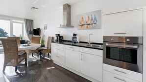 A modern kitchen with white cabinets, a built-in oven, and a dining area with wicker chairs and a round table. - Apartment with Sea View, Sauna, and High-Quality Furnishings (Sierksdorf)