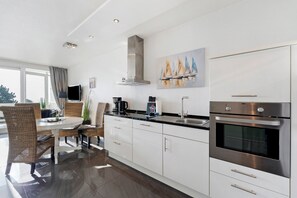 A modern kitchen with white cabinets, a built-in oven, and a dining area with wicker chairs and a round table. - Apartment with Sea View, Sauna, and High-Quality Furnishings (Sierksdorf)