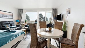 A modern hotel room with a bed, a dining table, wicker chairs, a TV, and a large window. - Apartment with Sea View, Sauna, and High-Quality Furnishings (Sierksdorf)