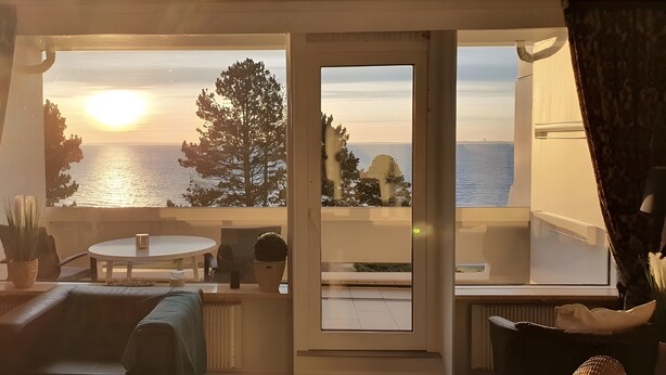 A room with a view of the sea at sunset, a balcony with a table and chairs, and a sofa. - Apartment with Sea View, Sauna, and High-Quality Furnishings (Sierksdorf)