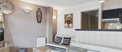 Een moderne woonkamer met een bank, salontafel en televisie. Er is een keuken met kastjes en een fornuis.