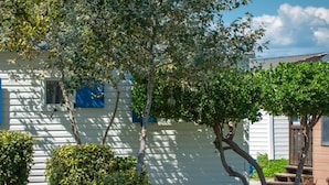 Une maison mobile blanche, dotée d’une petite fenêtre et d’un auvent bleu, entourée d’arbres et d’herbe.