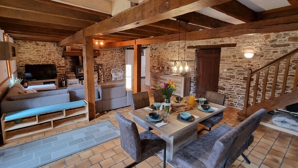 Un interior rústico con vigas de madera a la vista, paredes de piedra, una chimenea de piedra, un televisor y un comedor con mesa y sillas.