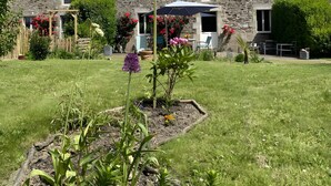 Una casa de piedra con techo inclinado, un jardín con diversas plantas y un cielo azul despejado.