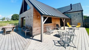 Une cabane en bois avec un toit en pente, une terrasse avec des meubles d’extérieur et, au fond, un bâtiment en pierre.