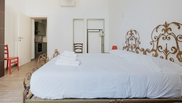 Un lit bien fait, avec du linge de lit blanc, une chaise rouge, un plancher en bois et une porte qui donne sur une cuisine.