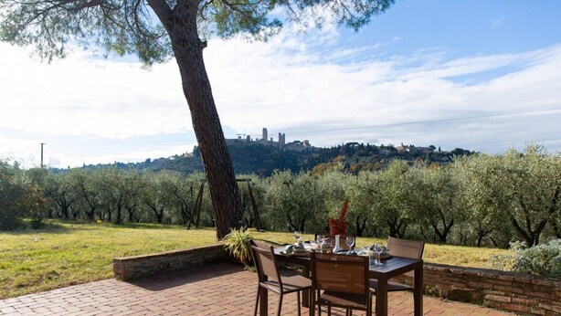 Un espace de restauration en plein air avec une table dressée pour deux personnes, entouré d’oliviers et offrant une vue sur le paysage urbain au loin.