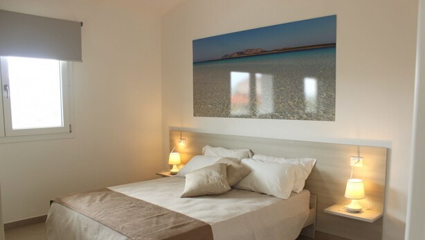 A bedroom with a bed, two bedside lamps, a window with a blind, and a painting of a beach scene on the wall. - Spacious 40 square meter apartment with independent access via magnetic card. The accommodation includes a fully equipped kitchenette, a living room with a comfortable sofa bed for two, a bedroom with a double bed, and a private bathroom with a shower an (Stintino)