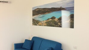 A room with a blue sofa, a wall-mounted air conditioner, and a framed coastal landscape painting. - Spacious 40 square meter apartment with independent access via magnetic card. The accommodation includes a fully equipped kitchenette, a living room with a comfortable sofa bed for two, a bedroom with a double bed, and a private bathroom with a shower an (Stintino)
