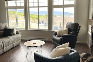 Ein Wohnzimmer mit einer Couch, einem Ledersessel und einem kleinen Tisch. Große Fenster bieten Blick auf einen Weinberg und Häuser.