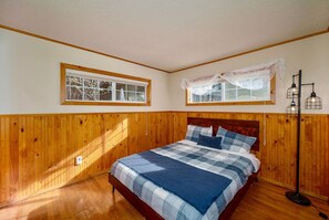 A bedroom with a wooden paneled wall, a bed with a blue and white checkered bedspread, two windows with white curtains, and a floor lamp. - The Blue Bear Cabin  3 Bedrooms, Hot Tub, (South Lake Tahoe)