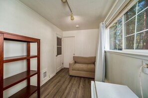 A room with a wooden shelf unit, a beige sofa, a window with a view of trees, and a door leading to another room. - The Blue Bear Cabin  3 Bedrooms, Hot Tub, (South Lake Tahoe)