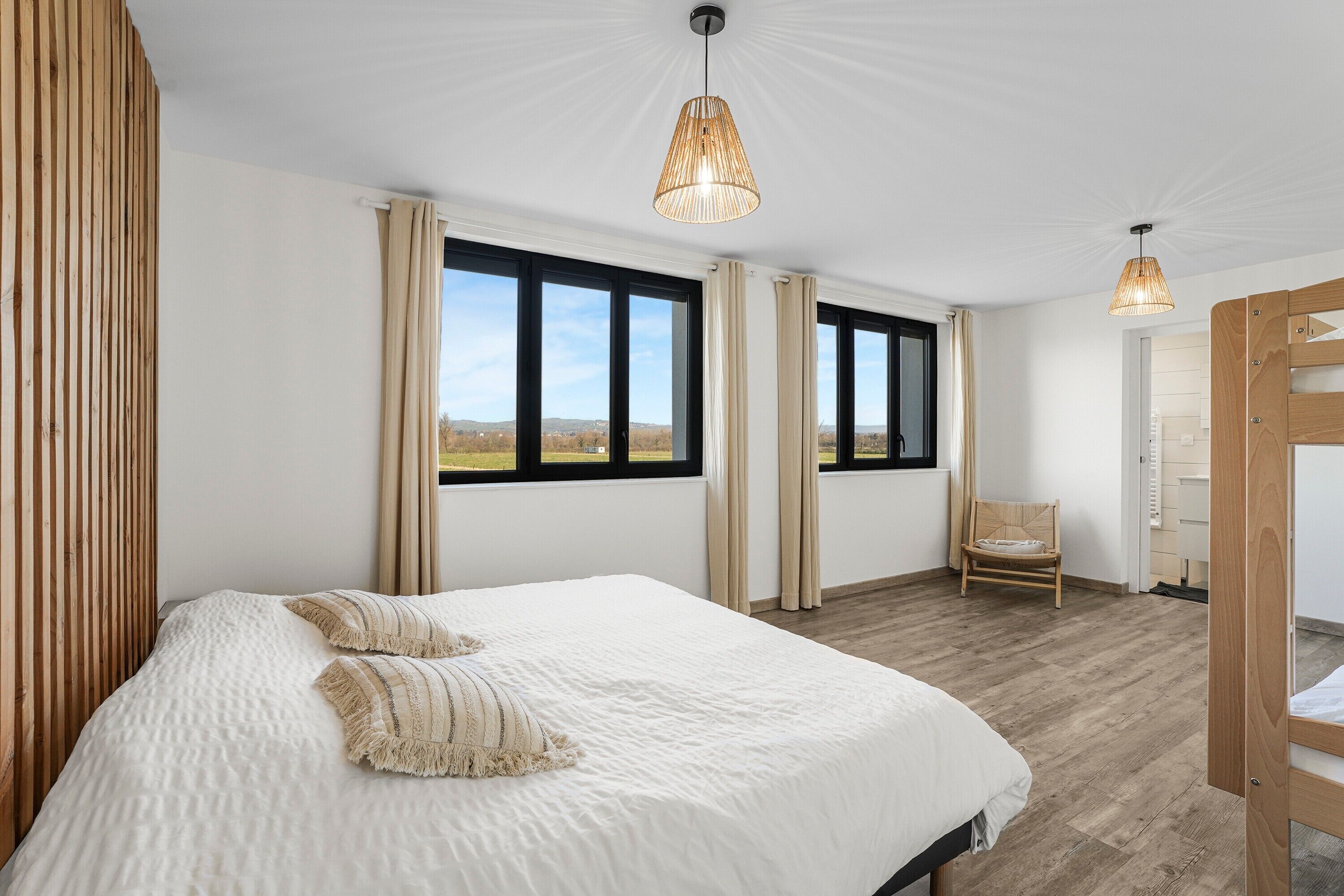 Ein Schlafzimmer mit einem großen Bett, zwei Fenstern, einem Sessel und einer Holztür.
