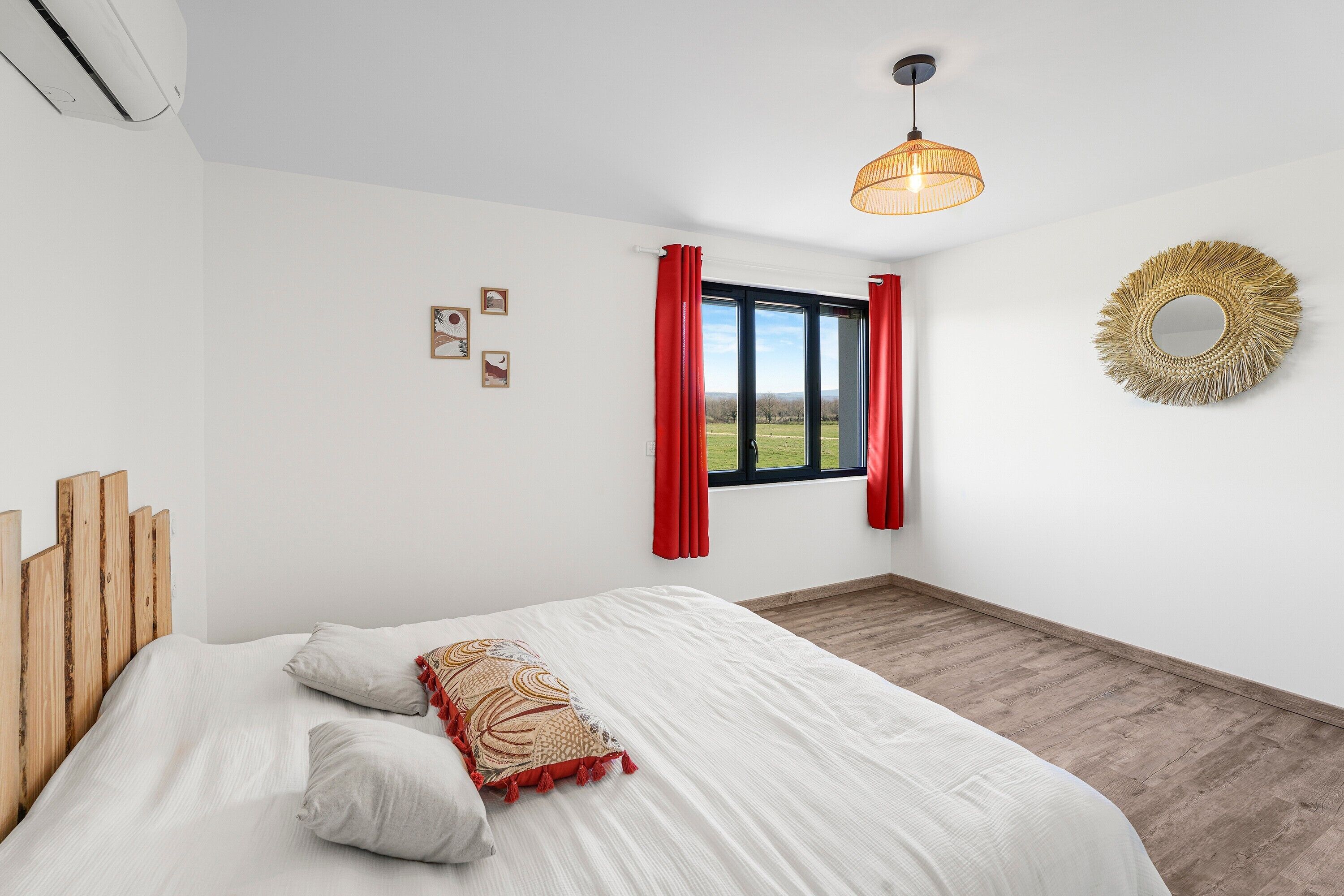 Una camera da letto con un letto, una finestra con tende rosse, uno specchio fissato al muro e un lampadario al soffitto.