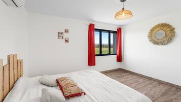 Una camera da letto con un letto, una finestra con tende rosse, uno specchio fissato al muro e un lampadario al soffitto.
