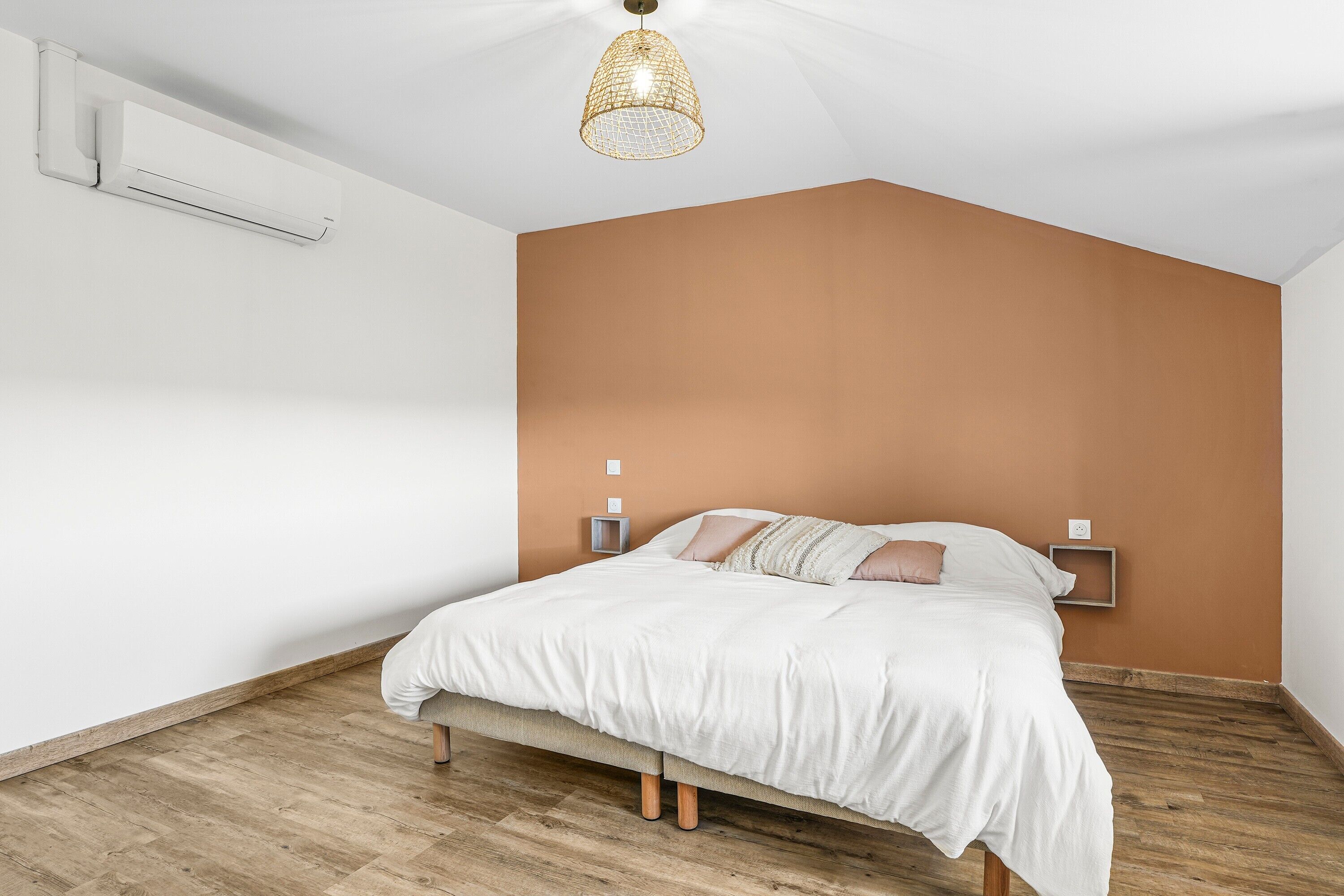 A bedroom with a bed, wooden flooring, a white wall, a brown accent wall, and a hanging light fixture.